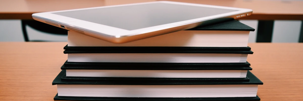 Realizziamo la manualistica che ti occorre, sia per la carta sia per il digitale Quattro libri rilegati posati su un banco scolastico. Sopra di essi, un tablet. Realizziamo la manualistica che ti occorre, sia per la carta sia per il digitale