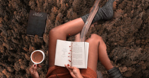 Dall'alto, una ragazza che legge un libro mentre con la sinistra tiene una tazza con cioccolata o caffè