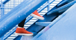 In un ambiente di scale dai toni blu, i piedi di un uomo con scarpe rosse che salgono